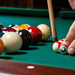 **Image Prompt:** A close-up of a billiards player meticulously chalking their cue, with a focus on the texture of the chalk and the concentration on the player's face. In the background, a slightly blurred billiards table with balls and cues. The lighting should be dramatic, highlighting the details of the chalking process.